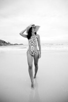 Black and white of young slender woman in stylish swimsuit and straw hat looking away while strolling on ocean beach