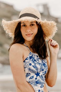 Young content female tourist in ornamental swimsuit touching hat while looking at camera on beach