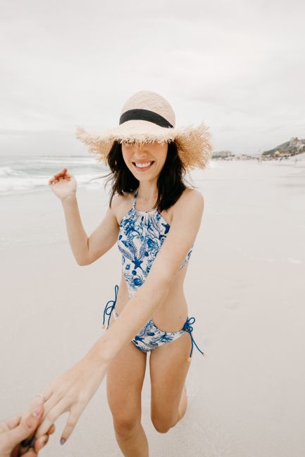 Young content female tourist in swimwear and straw hat strolling with crop unrecognizable partner on sandy ocean coast