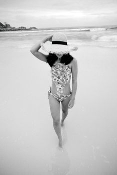 Black and white of anonymous female model in ornamental swimsuit touching straw hat while walking on sandy sea shore