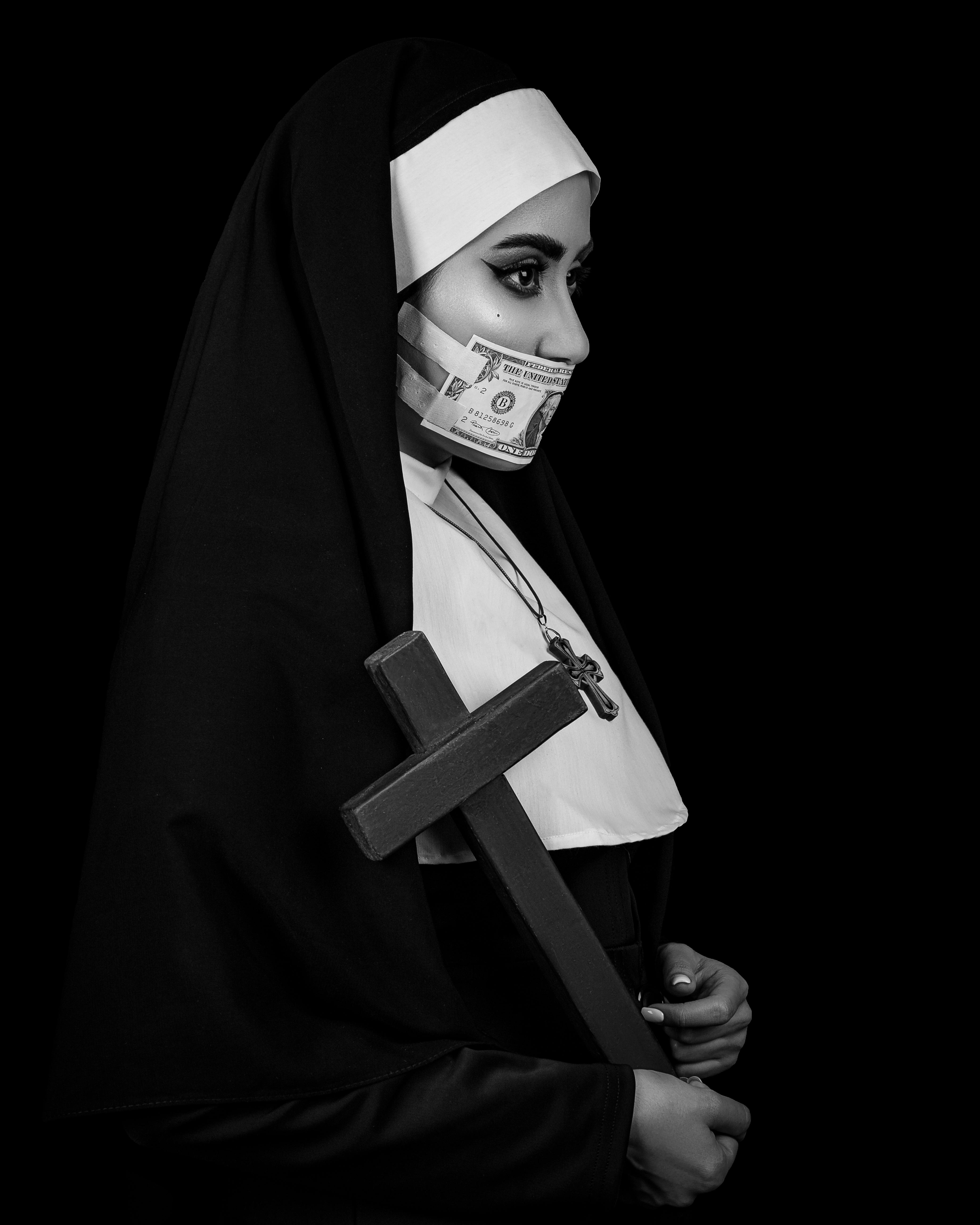 grayscale shot of a nun holding a cross with her mouth covered with a dollar cash