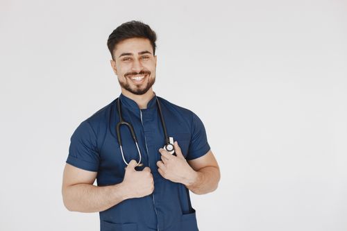 Free Portrait of a confident male doctor wearing a blue scrub suit and stethoscope, smiling in a studio setting. Stock Photo