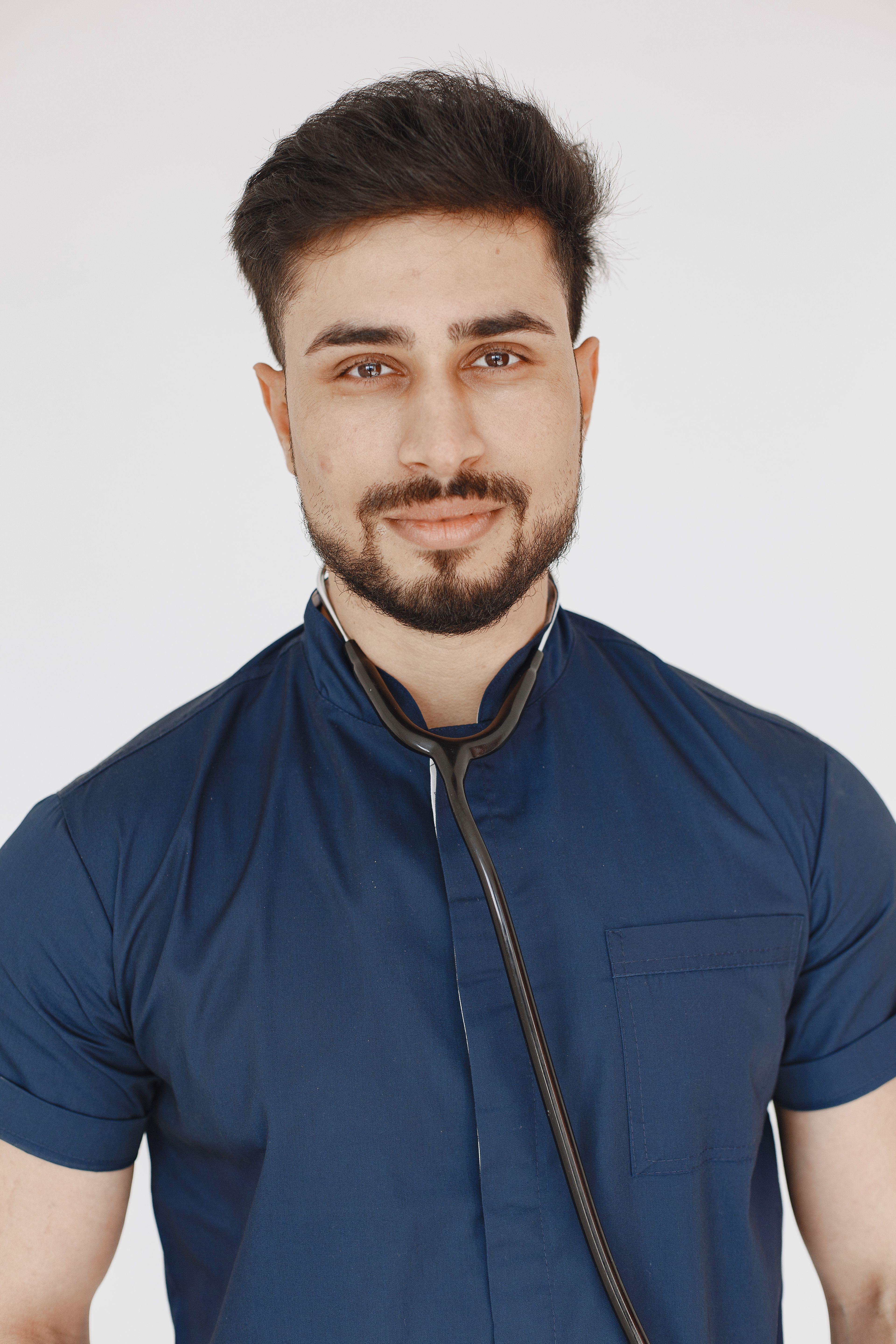 A bearded doctor smiling confidently in a studio setting, wearing a scrub suit and stethoscope.