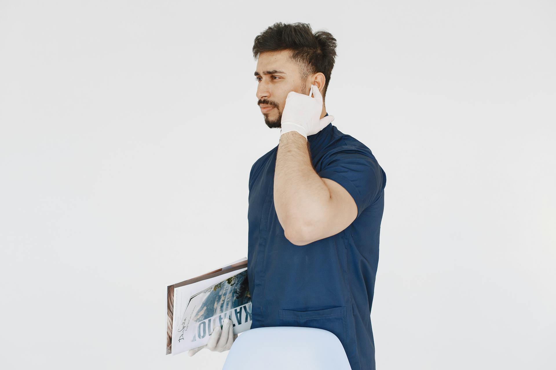 Bearded medical professional in scrubs, holding documents and wearing earpods in a studio setting.