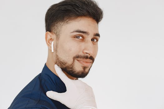 Young man with a beard wearing wireless earphones, smiling in a white setting with latex gloves adjusting his position.