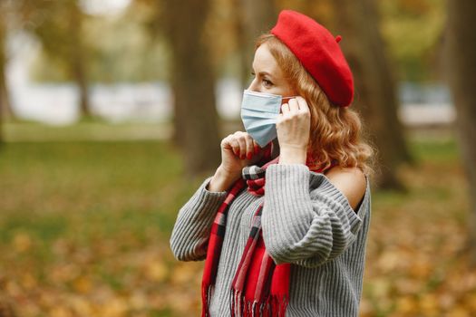 A woman in a red beret adjusts her face mask in a park during fall.