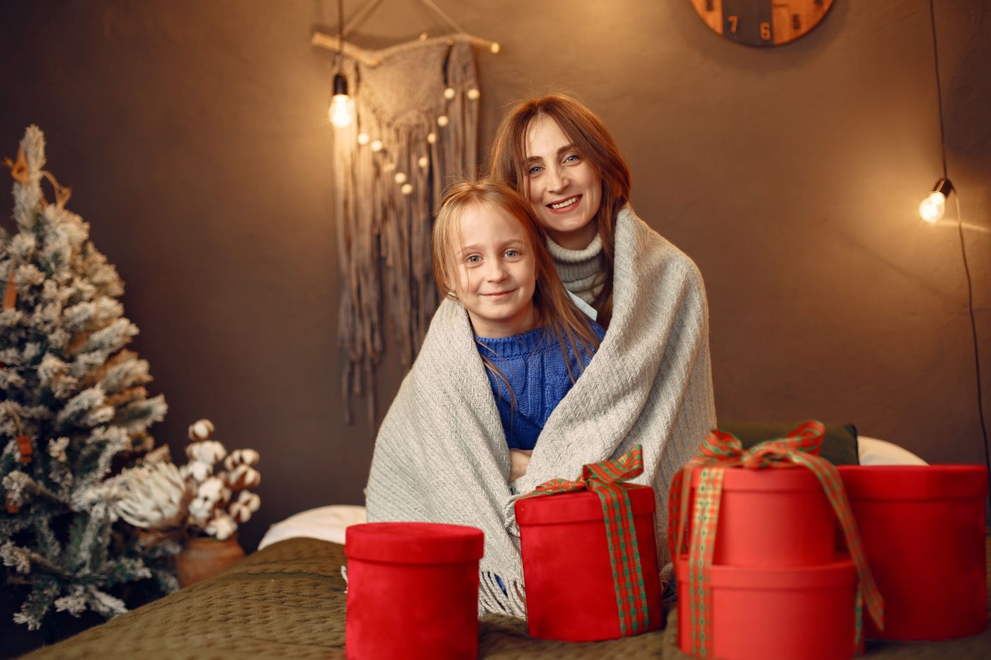 A mother and daughter wrapped in one blanket beside red gift boxes and string lights