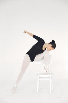 Ballet dancer in black leotard posing on chair against white backdrop.