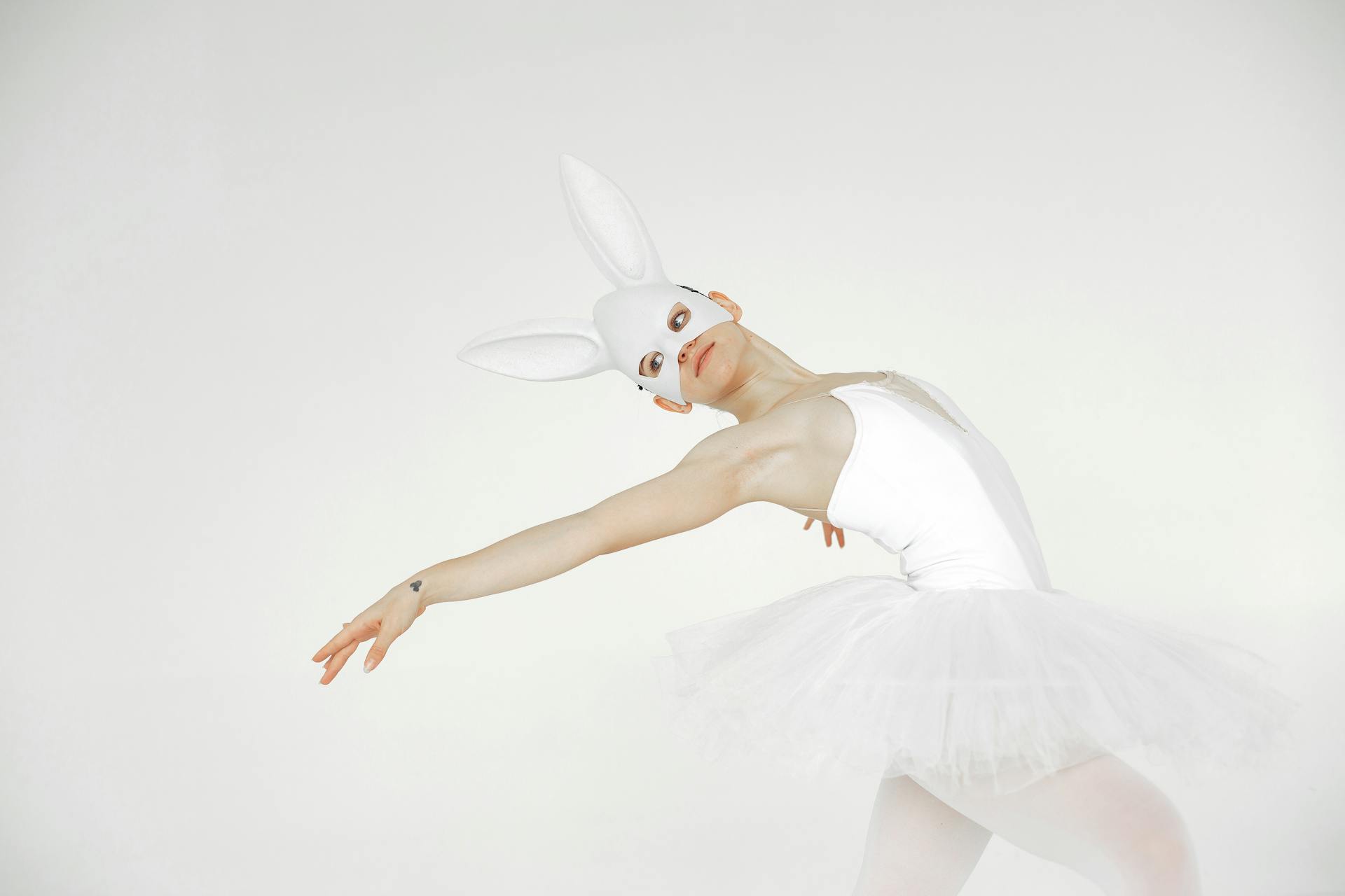 Ballet dancer in white tutu and rabbit mask performs graceful pose on white background.