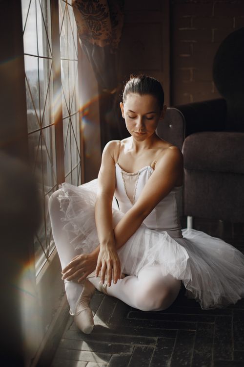 Free A graceful ballerina in a white dress sitting by a sunlit window, embodying elegance. Stock Photo