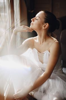 A graceful ballerina in a tutu, sitting by a window with eyes closed, basking in sunlight.
