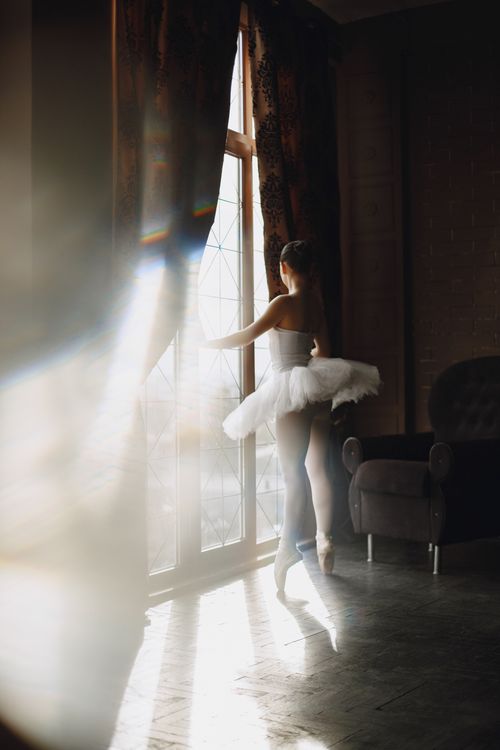 Free A ballerina poised elegantly by a window, illuminated by soft sunlight in a classic interior. Stock Photo