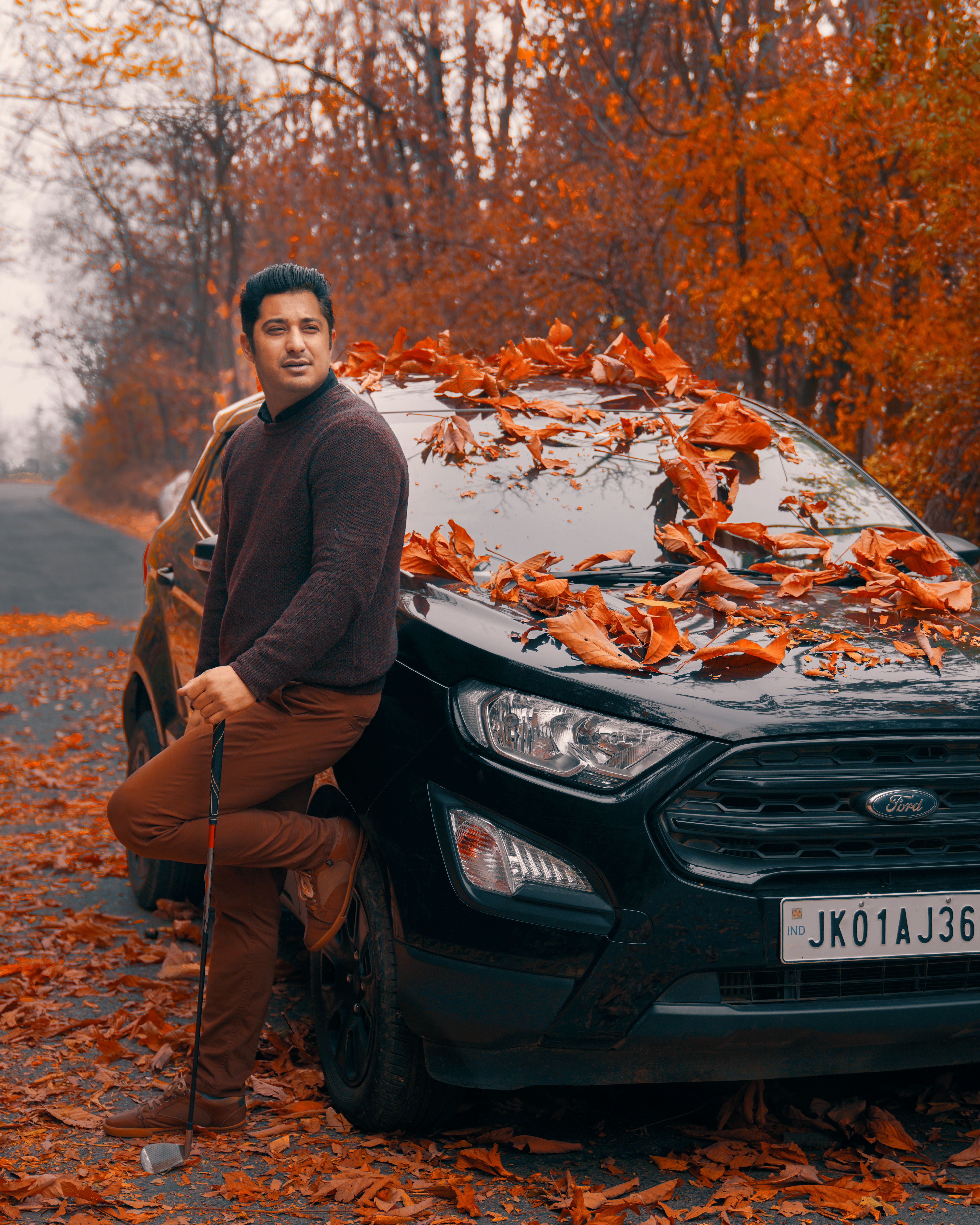 Man posing with black car covered in autumn leaves on a scenic road.