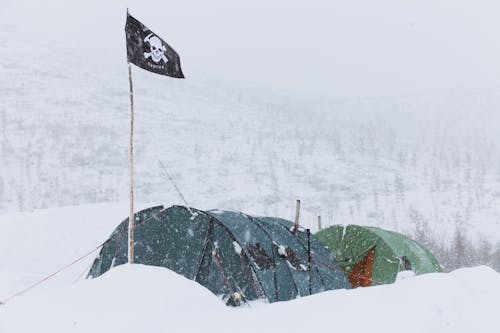 Colorful tents of expedition with flag with skull placed on ground in highland in snowstorm