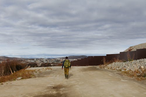 Back view of anonymous male with backpack walking in outskirts of city under cloudy sky