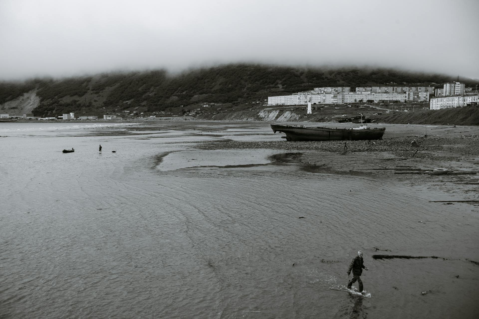 A serene, foggy coastal scene with a shipwreck and distant buildings under overcast skies.
