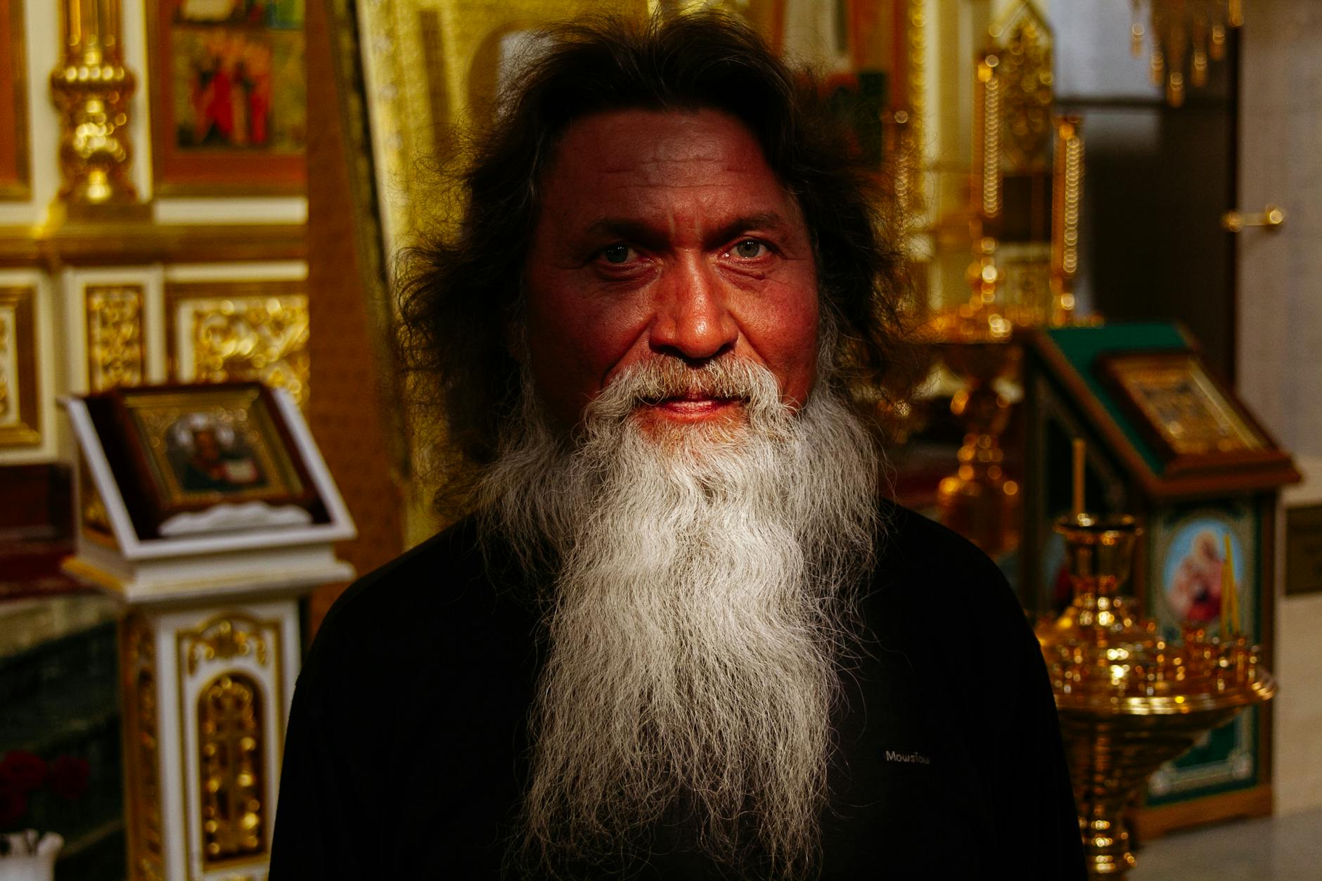 Peaceful senior Orthodox priest with gray beard in cassock standing in church near golden icons and frescoes and looking at camera