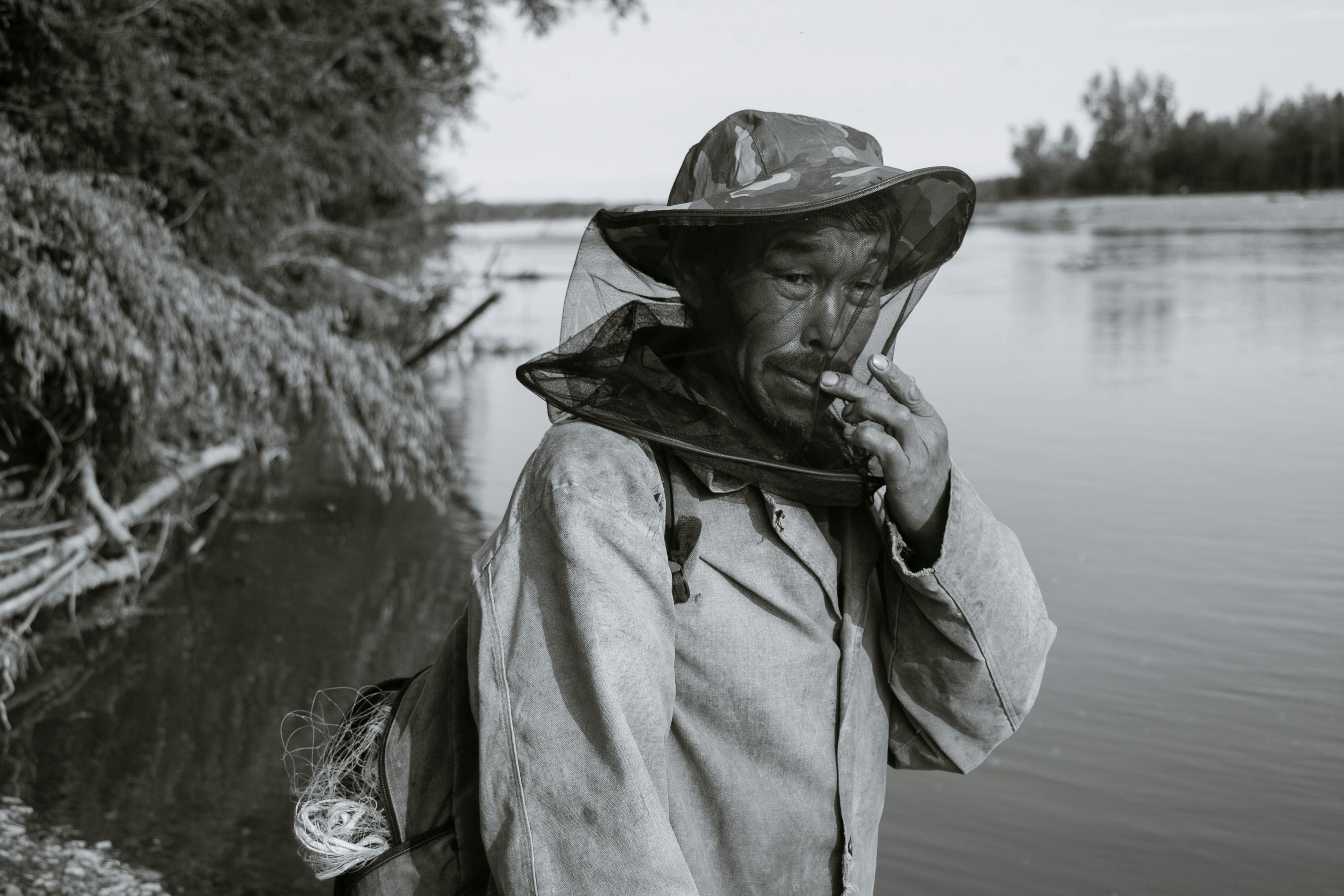 Side view of black and white middle aged Asian male in casual clothes and anti mosquito head net hat standing on lake shore and looking at camera before fishing