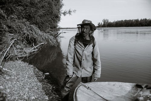 Free Black and white of calm mature ethnic fisherman in anti mosquito head net hat standing in shallow water of lake near wooden boat and looking at camera Stock Photo