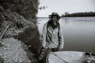 Black and white of calm mature ethnic fisherman in anti mosquito head net hat standing in shallow water of lake near wooden boat and looking at camera
