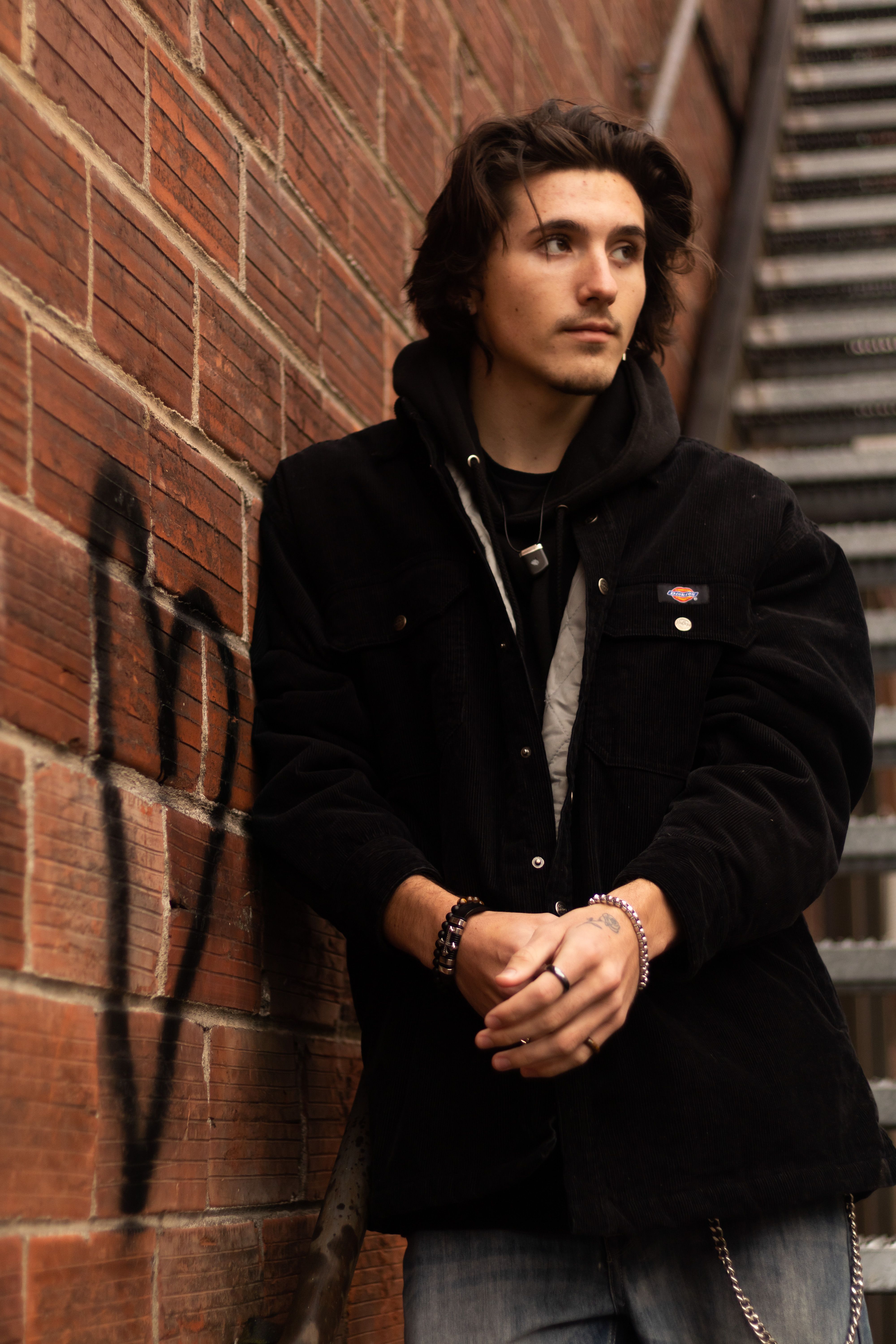 Young man in black hoodie posing against a brick wall with heart graffiti.