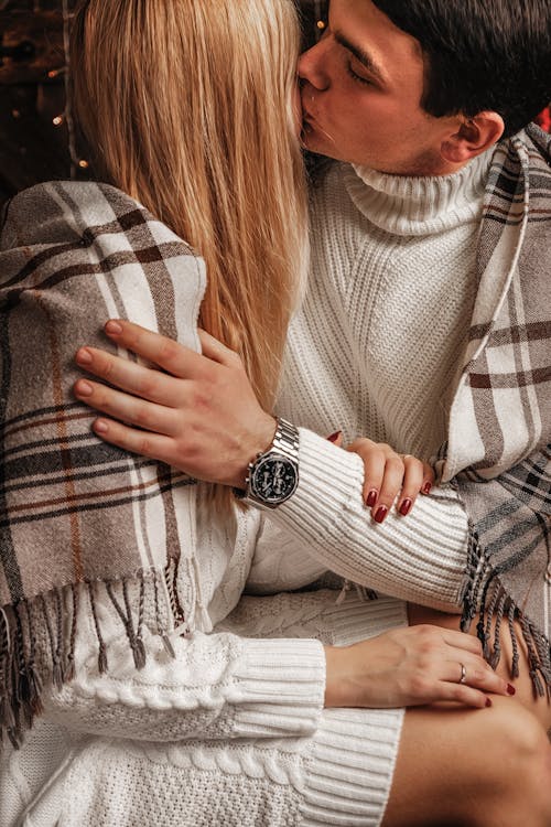 Crop young couple wearing warm sweaters covering shoulders with scarf cuddling and kissing tenderly at home