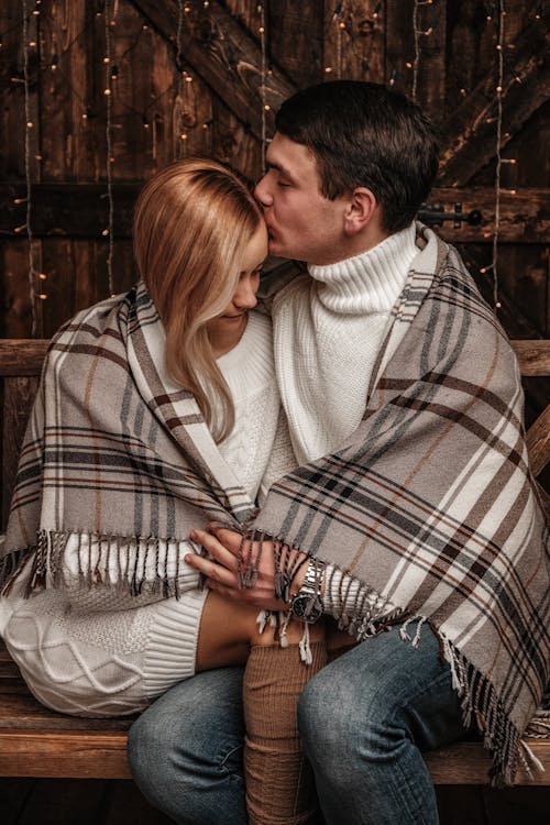 Loving young man hugging and kissing beautiful girlfriend forehead while sitting together on wooden bench under cozy plaid