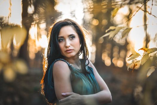 Young charming female in green dress with makeup and long hair standing in park and looking away