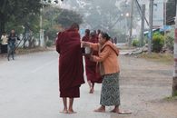 A woman offers alms to a Buddhist monk on a street in Shan, Myanmar, showcasing cultural traditions.