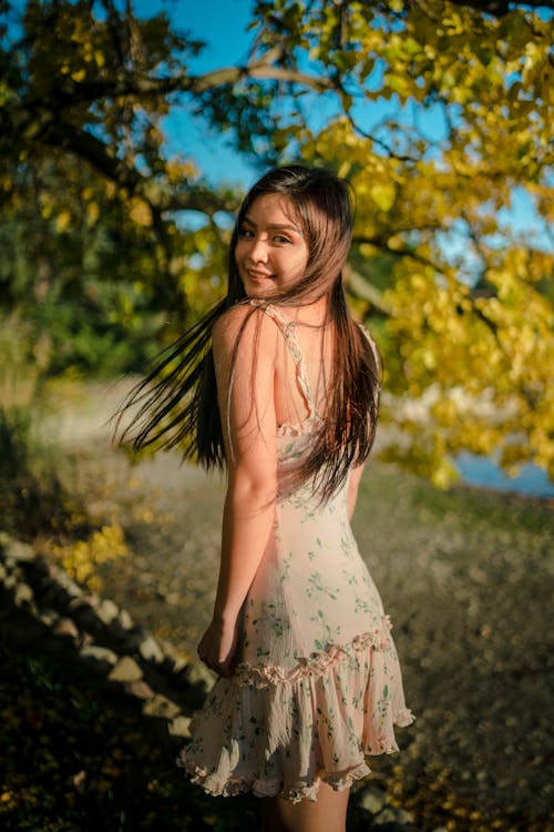 Charming young female in dress with long hair standing near tree and looking at camera smiling