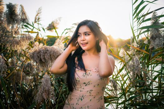 Happy Asian female with long dark hair in summer outfit touching hair and standing in tall green plants in nature in sunny day