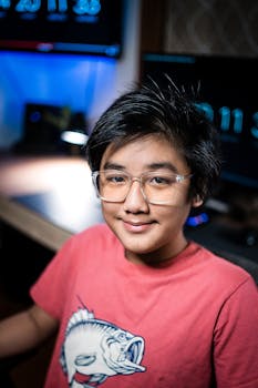 Smiling Asian boy wearing glasses and red t-shirt, sitting indoors with computer screens.