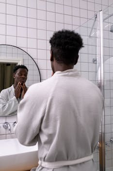 An adult man in a white bathrobe examines his face in a modern bathroom mirror.