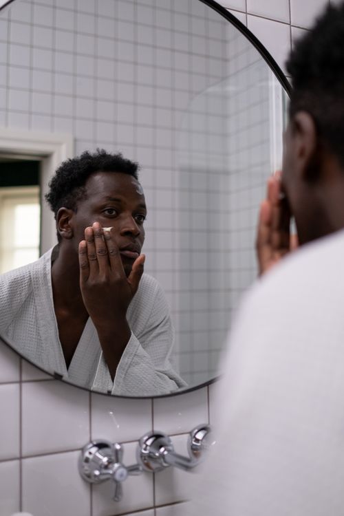 Free African American man applying facial cream while looking in a bathroom mirror, promoting skincare routine. Stock Photo