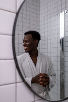 A man in a bathrobe smiles while looking sideways in a bathroom mirror.