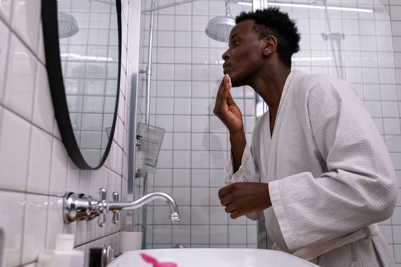 A Black man in a white robe leans toward his bathroom mirror, pressing a cotton pad to his jaw while checking his skin.