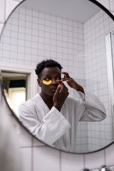 A man in a bathrobe applies under-eye masks while looking in the mirror.