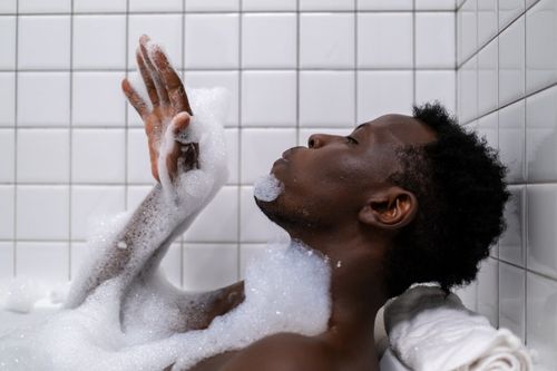 Free Side view of an adult relaxing in a bathtub with bubbles against a white tiled background. Stock Photo