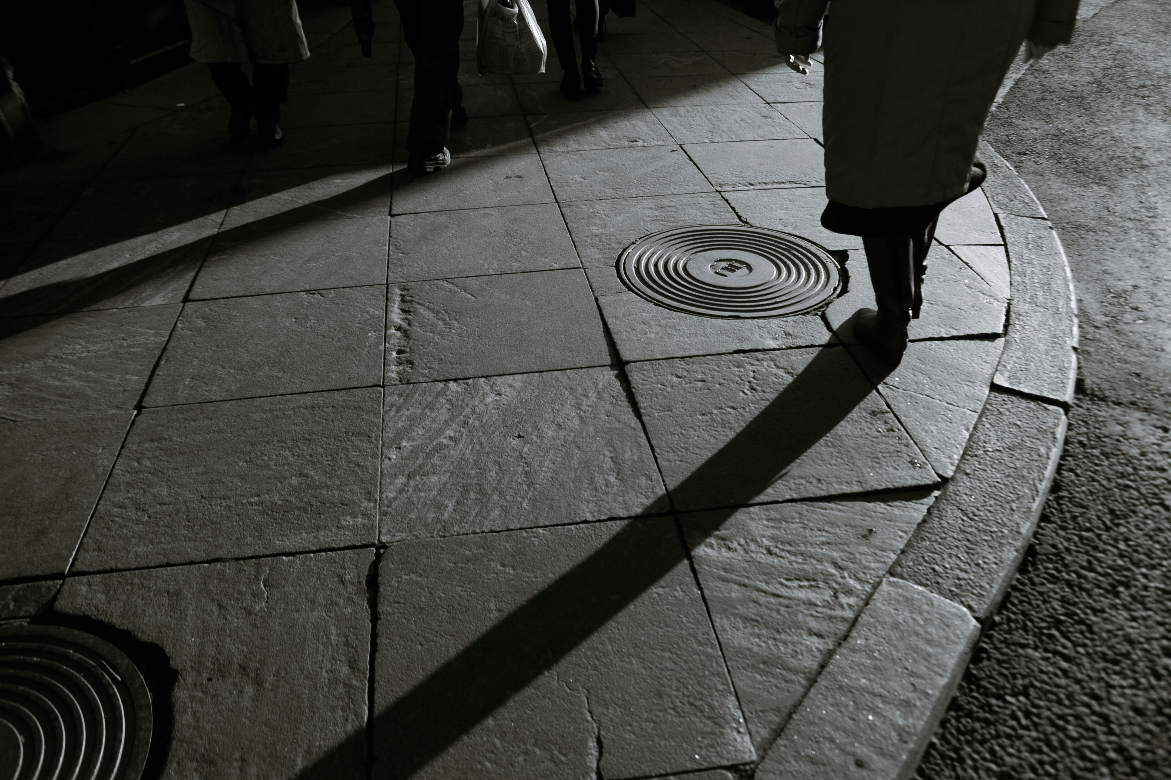 Free High-angle black and white shot of pedestrians casting long shadows on city pavement at dusk. Stock Photo