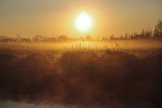 Rising morning sun shining over dry grass and rippling pond in mist in countryside