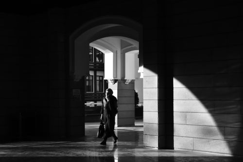 Free A woman walks through a sunlit archway, contrasting shadows in black and white. Stock Photo