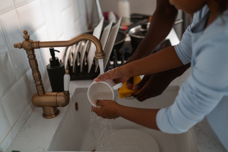 A child and adult washing dishes together at a brass kitchen sink, promoting teamwork and family chores.