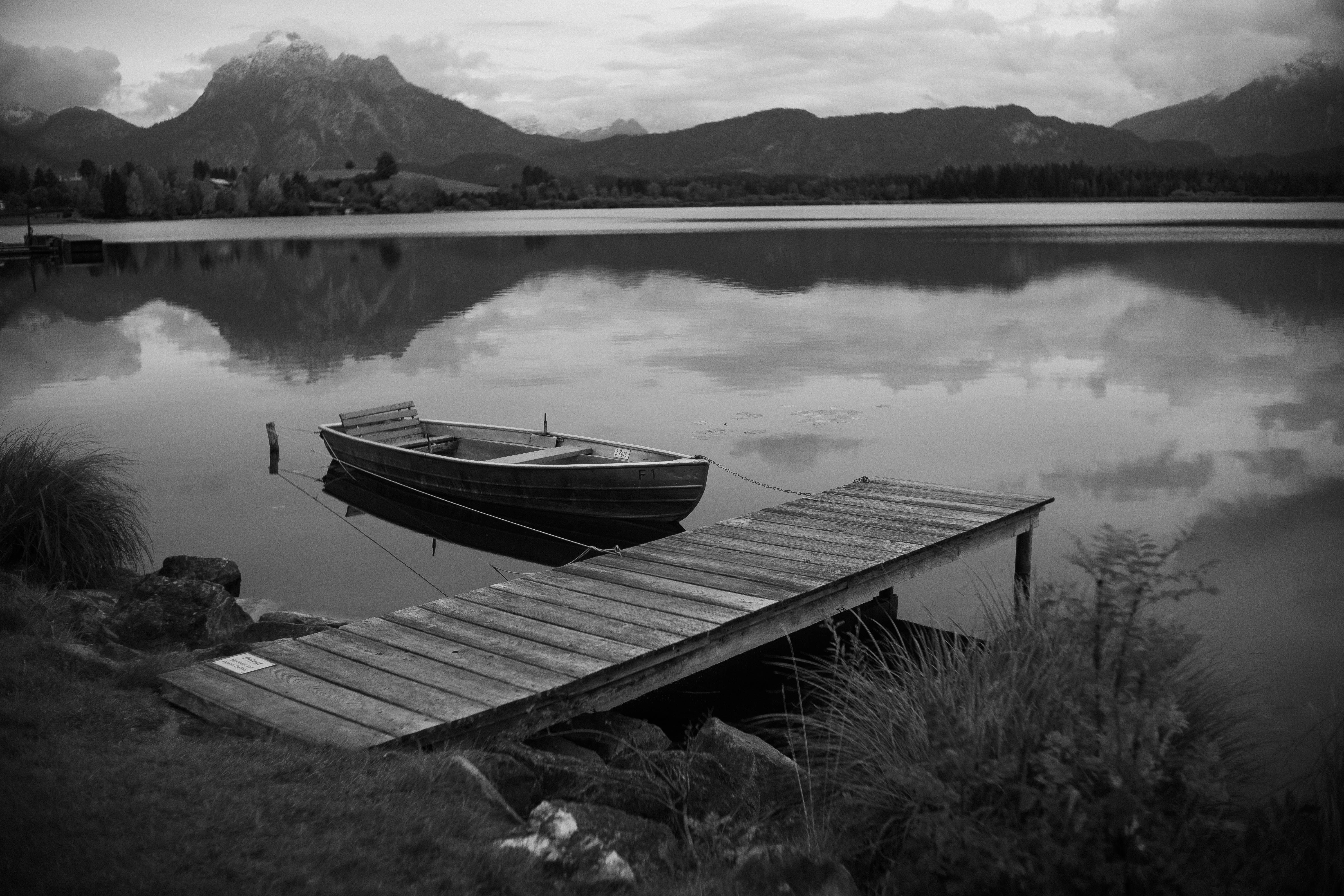 Serene black and white image of a lake with a wooden boat and dock, reflecting mountains.