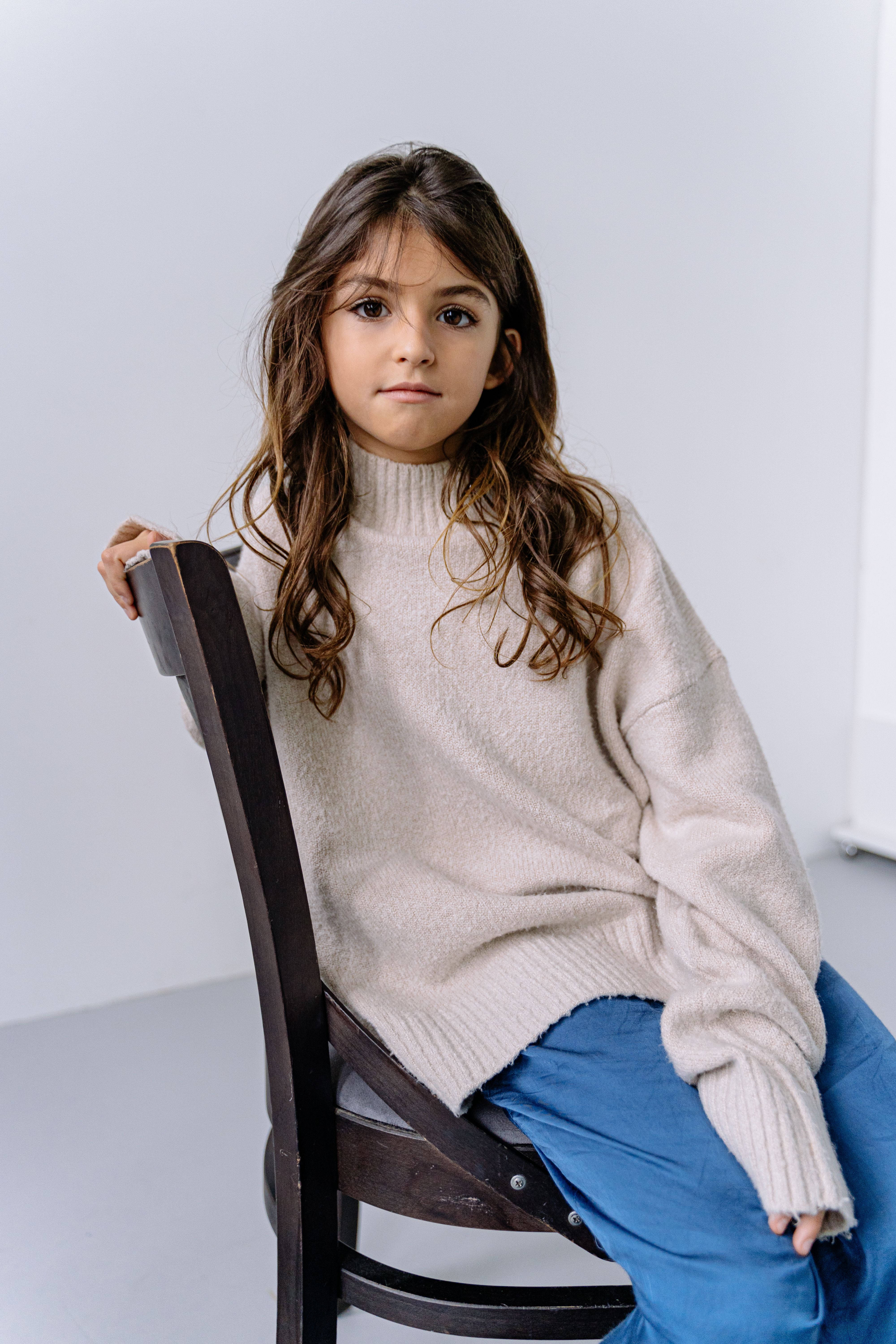 Young girl sitting on a wooden chair wearing a cozy sweater. Neutral background.