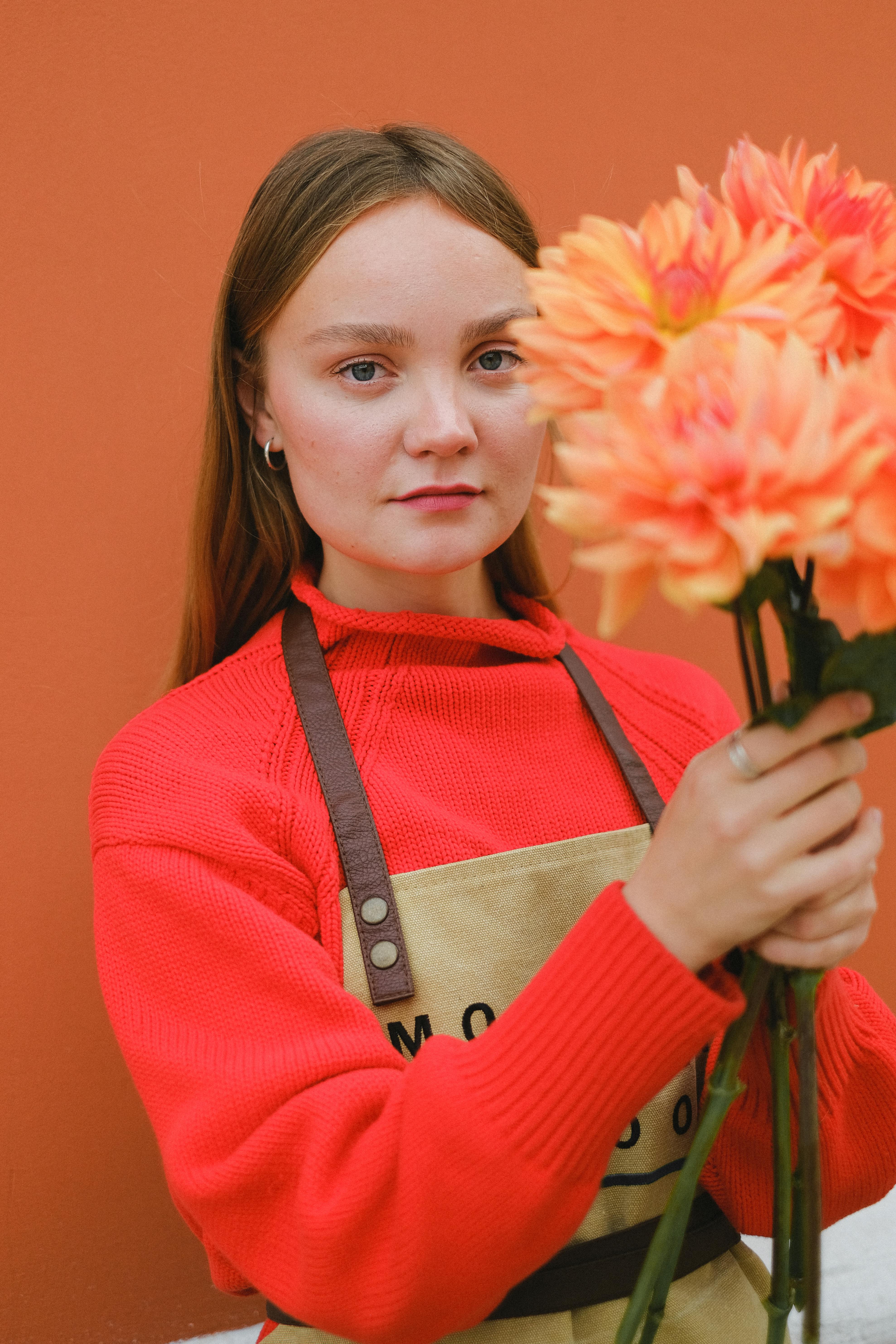 Portrait of a female florist holding orange dahlias against a matching background, blending artfully with her red sweater.