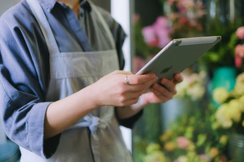 Free Florist in apron using digital tablet inside flower shop with blurred floral background. Stock Photo