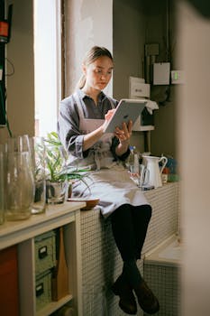 A florist wearing an apron sits indoors using a digital tablet in a cozy workspace with plants.