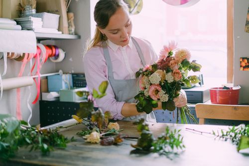 Free Florist carefully arranging a bouquet with vibrant flowers and leaves indoors. Stock Photo