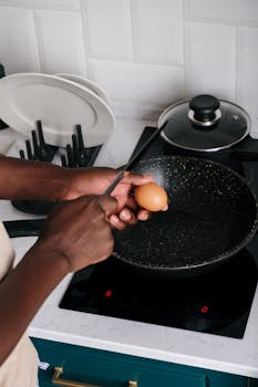 Close-up of hands cracking an egg into a frying pan on an electric stove.