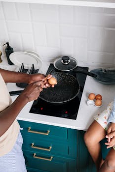 A person cracking an egg into a frying pan on an electric stove, ready for breakfast.